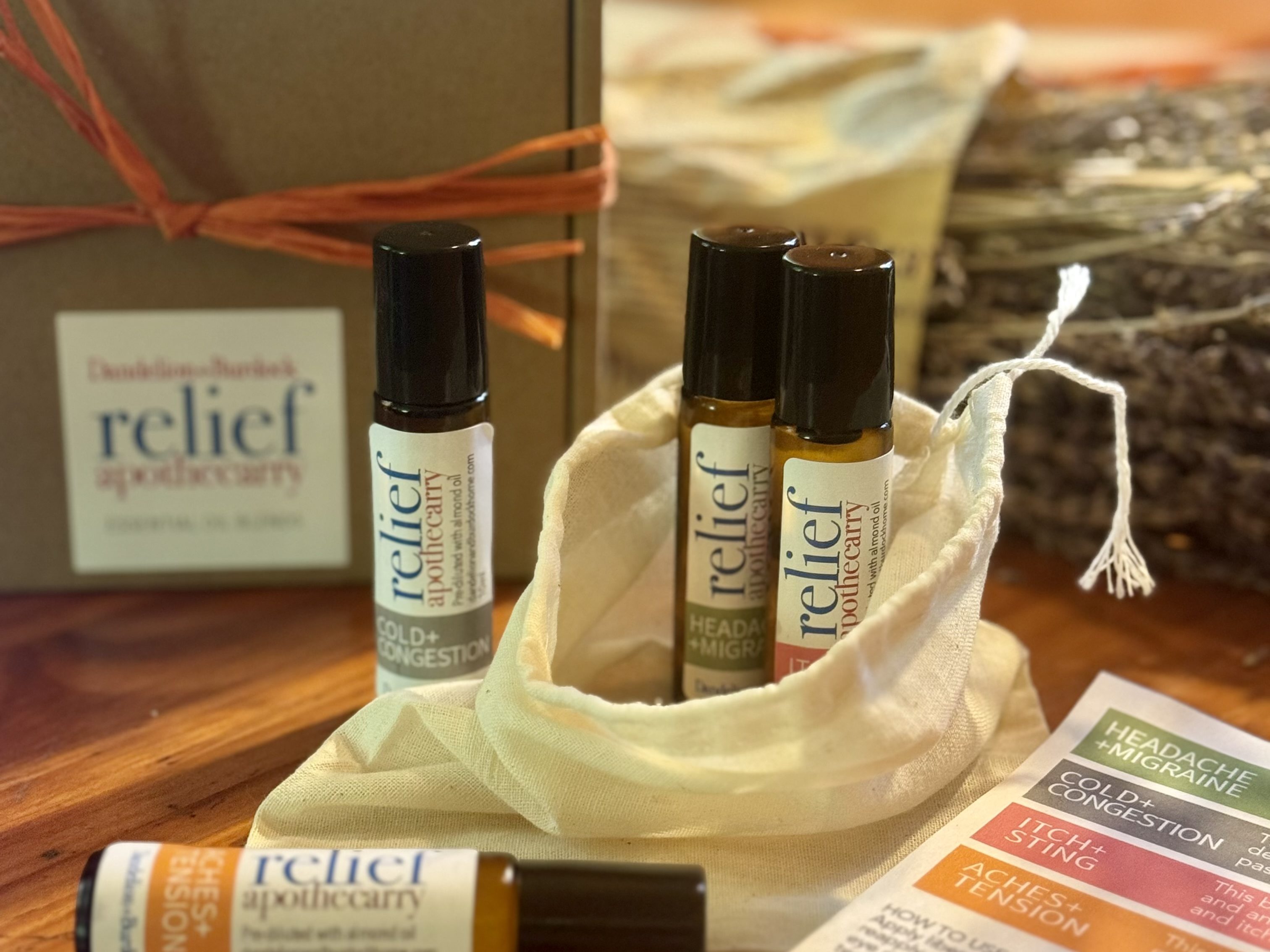 A collection of four essential oil roll-ons from the Dandelion+Burdock Relief Apothecary Set. The roll-ons are placed in a natural fabric pouch, with a greeting card detailing their uses, sitting on a wooden table.